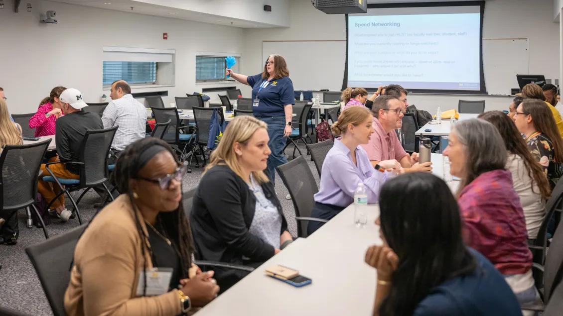 People sitting at tables and talking in a classroom with an instructor giving a presentation