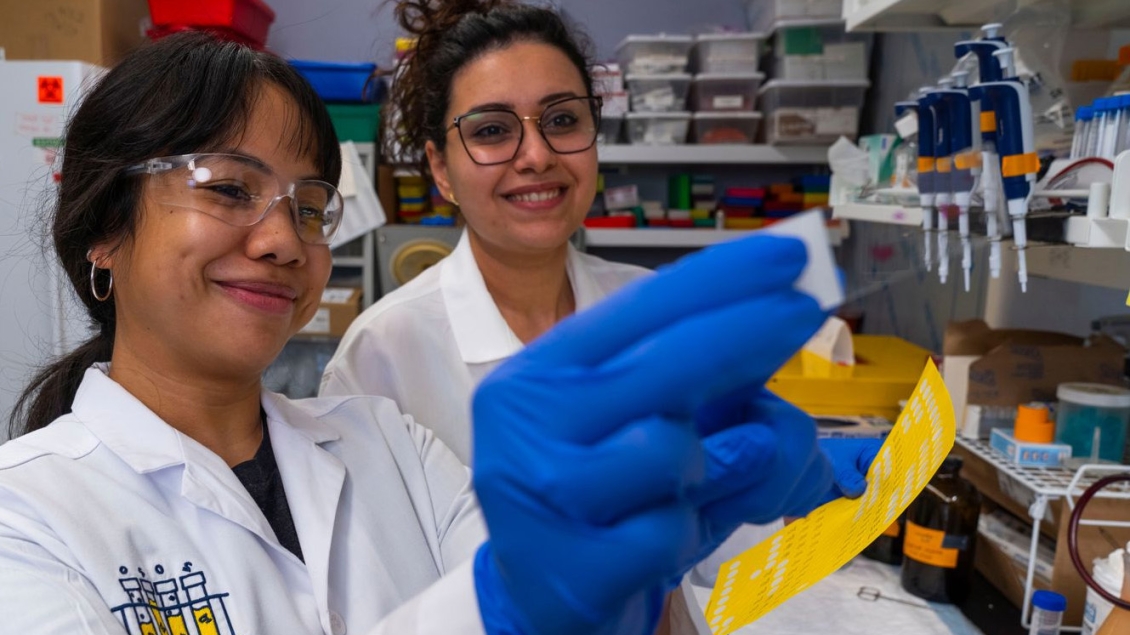 Two lab workers in white coats examine a small item held up by one of them. They are surrounded by shelves of lab supplies and large equipment is visible in the background.