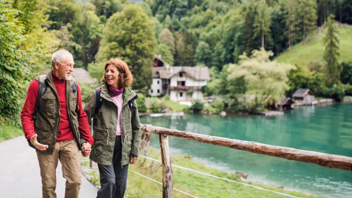 A man with white hair wearing a red shirt and green vest smiles, walking hand in hand with a woman with red shoulder-length hair, wering a purple scarf and green jacket. They are walking along a scenic river with trees in the background.