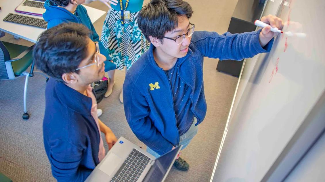 Two students write on a white board and hold a laptop
