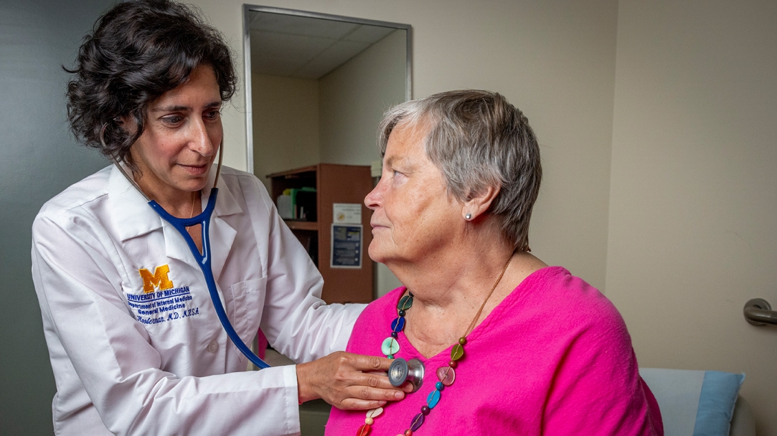 Dr. Hooberman, wearing a University of Michigan lab coat, uses a stethoscope to listen to the chest of a patient in a bright pink top and colorful beaded necklace during a clinical exam in a medical office.