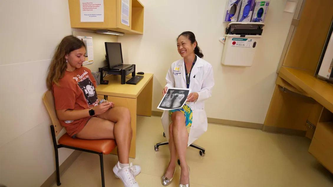 A girl sits in a chair as a doctor reviews her x-ray images.