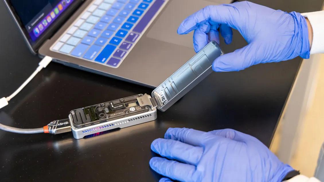 A researcher wearing blue gloves loads a small sequencing device connected to a laptop.