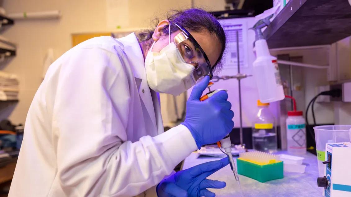 Scientist wearing goggles, mask, and gloves uses a pipette while working at a lab bench.