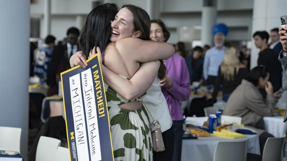 Two people hug in a crowded indoor celebration space while one holds a maize-and-blue sign reading “I MATCHED! into Internal Medicine at MICHIGAN!” with tables and other attendees in the background.
