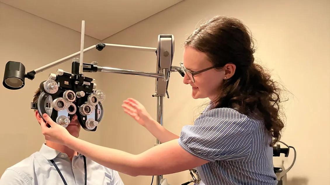 Resident adjusts a phoropter during an eye exam while another resident looks through the device, demonstrating clinical training in vision testing.