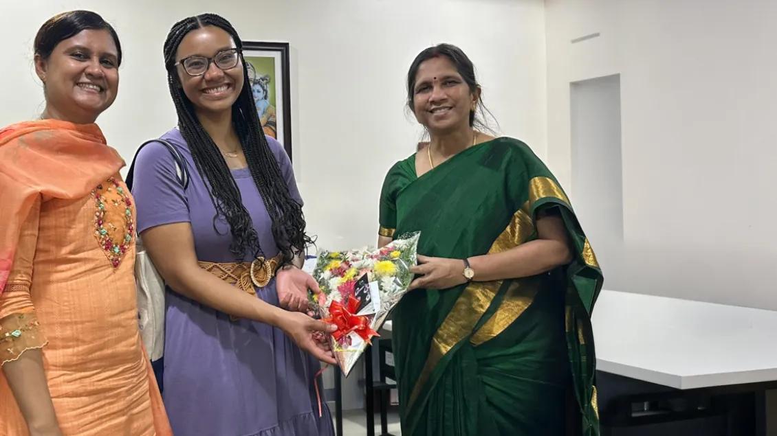 Otana Jakpor, MD, receiving a floral bouquet at Aravind. Two Aravind staff members in an orange and dark green sari stand on either side of her. 