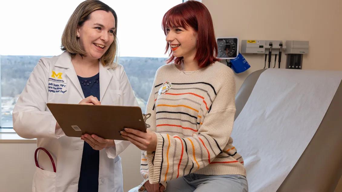A doctor in a white coat stands next to a patient wearing a striped sweater who is seated on an exam table. They are both smiling and holding a clipboard together. The doctor is writing something on it.