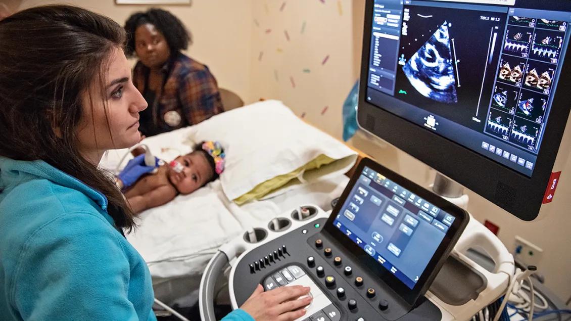 A medical professional performing a cardiovascular imaging exam on a baby who is lying on a bed in the background. There is a monitor and device controls in front of the staff, and the baby's mom is seated next to the bed.