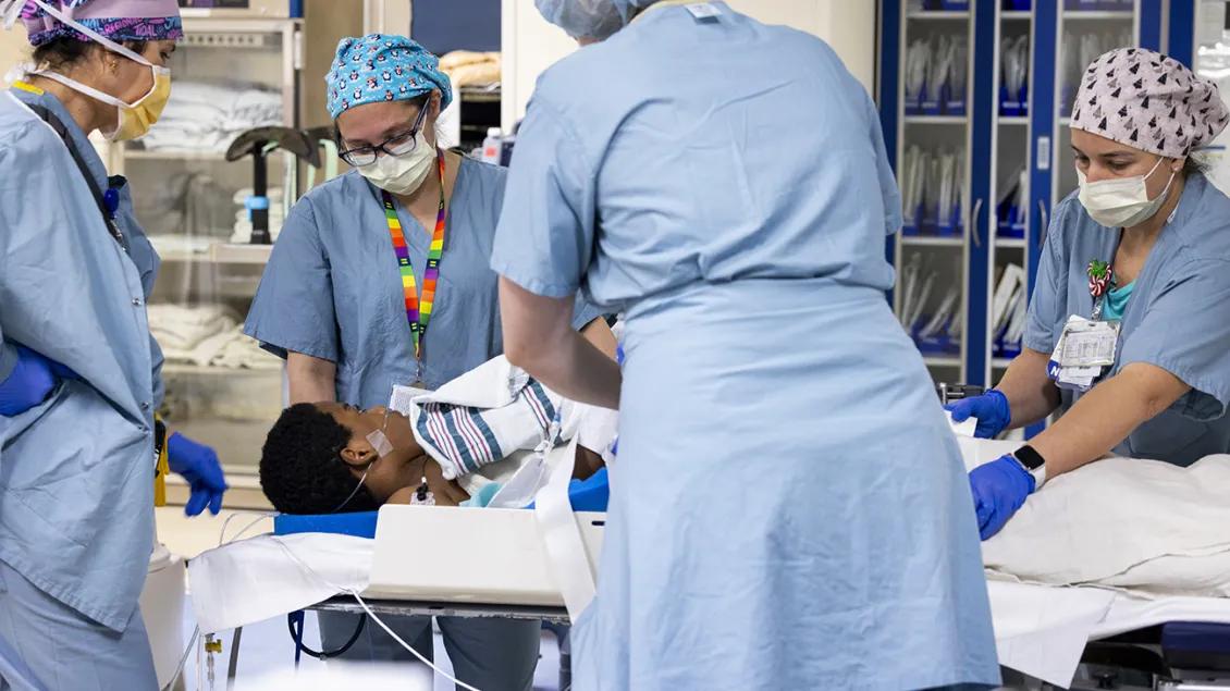 Four medical professionals wearing masks, surgical caps, and scrubs stand around a child who is lying on a bed and covered with blankets. There are shelves of blankets and unidentifiable medical equipment in the background.