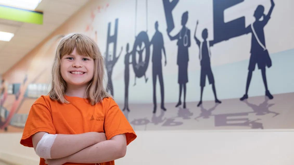 A young patient wearing an orange t-shirt and a bandage around her arm smiles and stands with her arms crossed in front of a wall mural that spells out "hope."