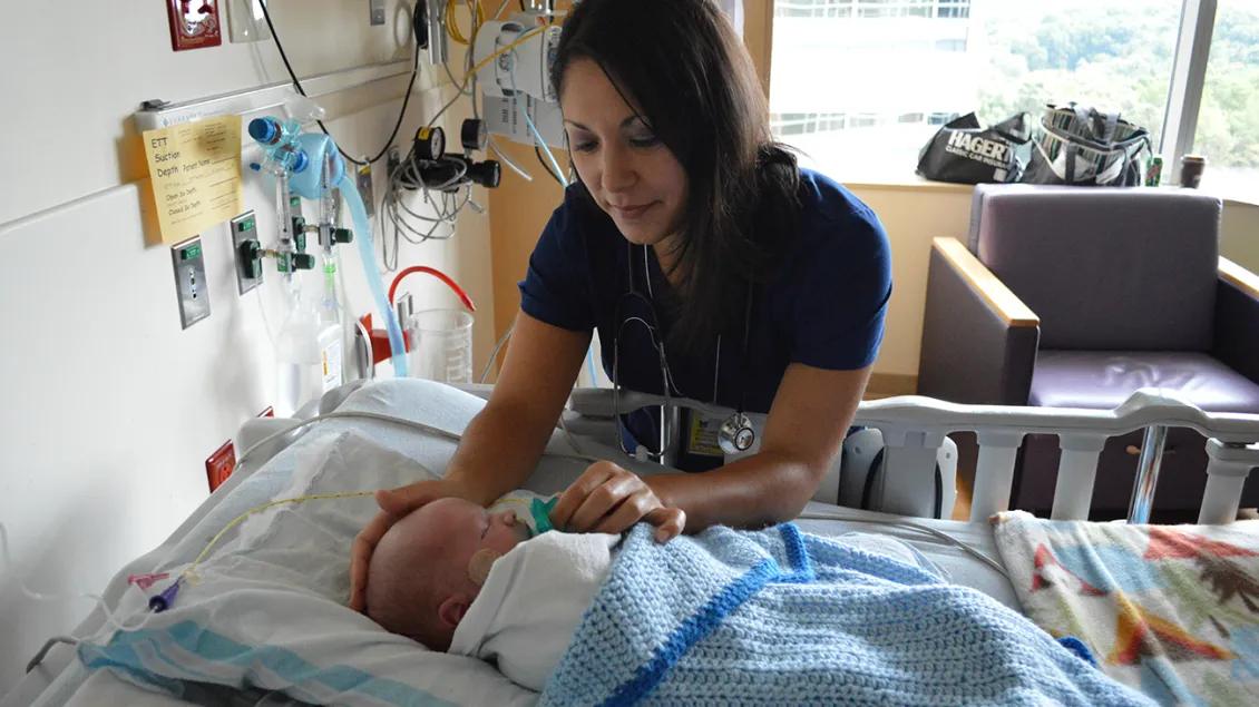 A medical professional wearing blue scrubs leans over a warmly covered baby who is sleeping in a bed. The professional is gently placing a hand on the baby's head as she puts a pacifier in the baby's mouth.