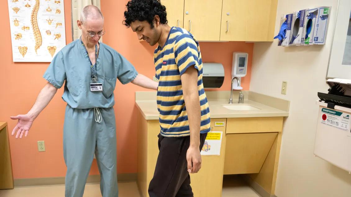A doctor wearing blue scrubs stands in an exam room with a young man who is wearing a striped shirt. The young man appears to be stepping intentionally as if they are doing a movement assessment.