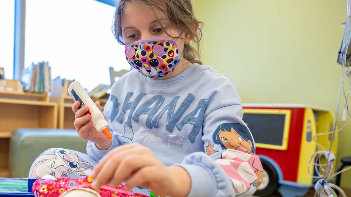 A young girl wearing a mask and a blue sweatshirt appears to be crafting, gluing something on to a doll's dress.