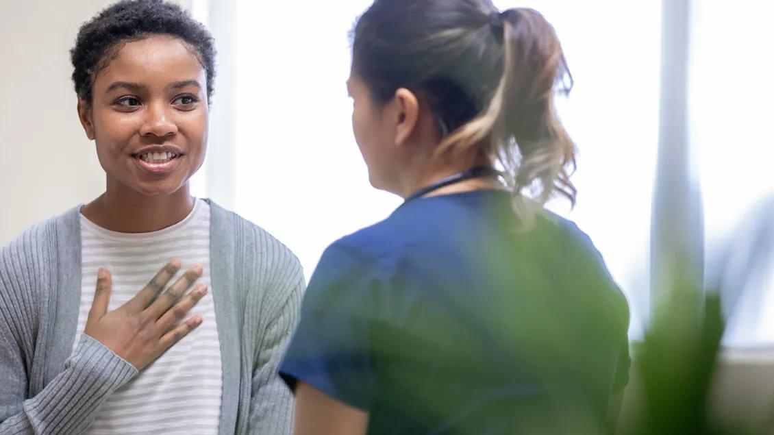 A young adult patient, smiling and placing her hand over her chest in an emotional way, talks with a medical professional who is wearing blue scrubs.
