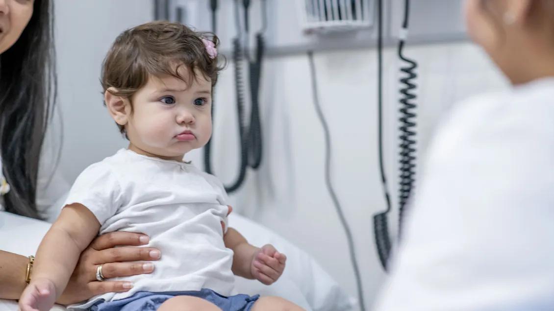 A baby with short curly hair sits on an exam table. She is held up by a woman on the left who is just visible, and a blurred medical professional smiles at her from the right.