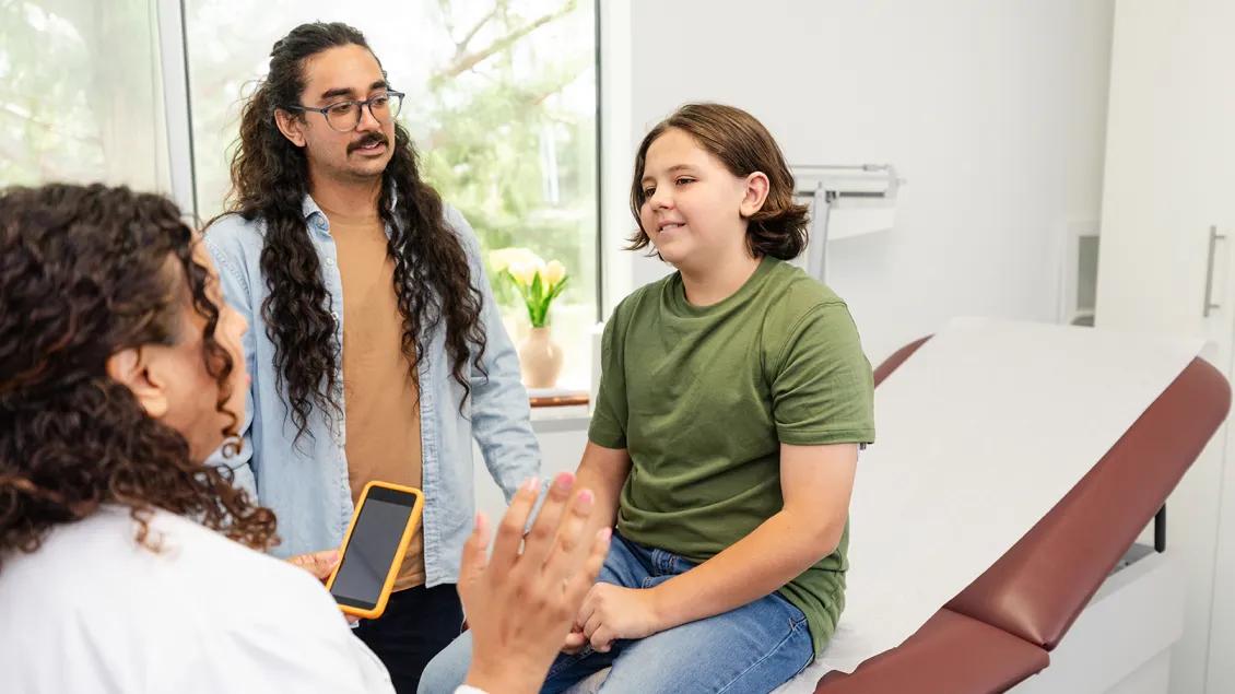 A young patient seated on an exam table and a caregiver talk with a doctor.