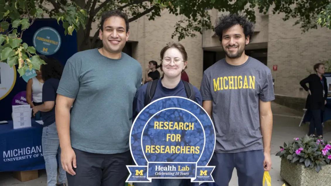 three smiling scientists in front of a sign stating Research for Researchers