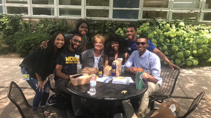 Group of Black and brown-skinned medical students with white woman seated at a table outside with flowering bushes in background