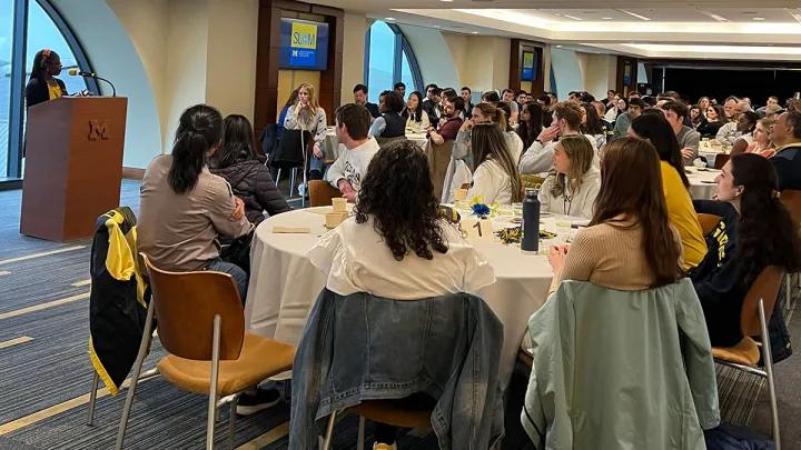 Group of students seated at round tables looking at Black woman standing at podium