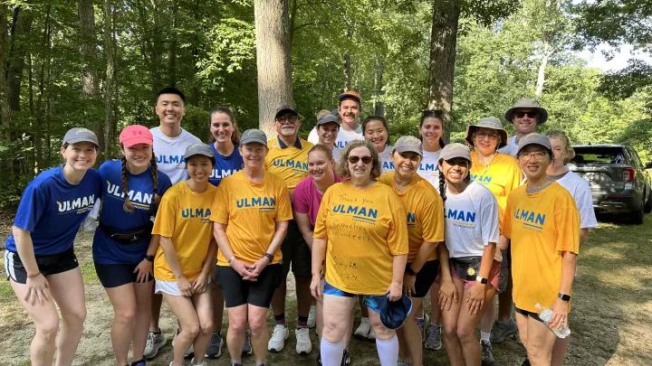 A group of 19 Cancer to 5K participants wearing a mix of maize, blue and white shirts pose outdoors in front of trees