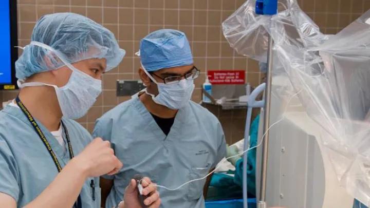 Two medical professionals wearing blue surgical scrubs, caps, and masks stand in a hospital or surgical room. One person is holding a medical device connected to a larger machine, which is partially covered with clear plastic. The background includes tiled walls, medical equipment, and supplies.