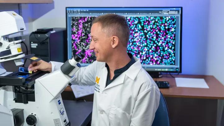 Dr. Sebastian Werneburg in front of a large microscope with an colorful research image on a monitor behind him