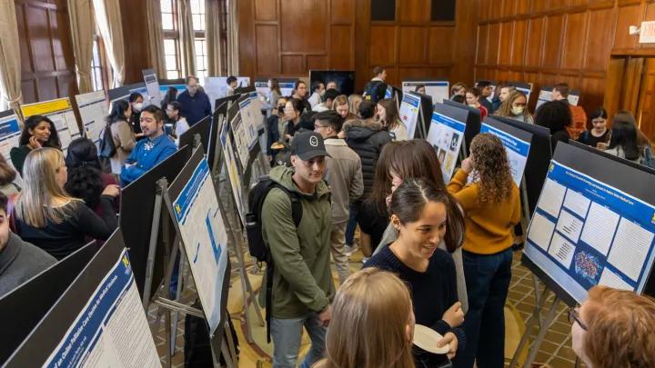 Attendees mingle and examine research posters displayed on easels in a wood-paneled room during an academic poster session or conference.