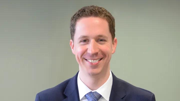 Headshot of a smiling man with short brown hair wearing a navy suit, white shirt, and blue patterned tie against a plain gray-green background.