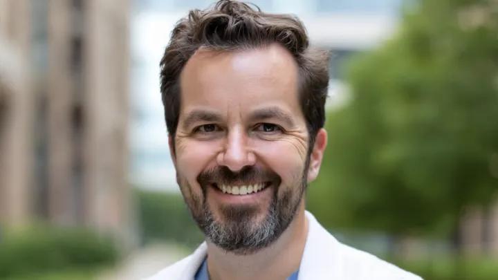 Headshot of a smiling man with short brown hair and a salt-and-pepper beard, wearing a white coat outdoors against a softly blurred background of trees and buildings.
