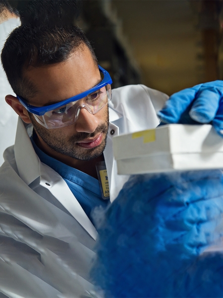 Brown-skinned man wearing goggles and blue gloves examining something in white box