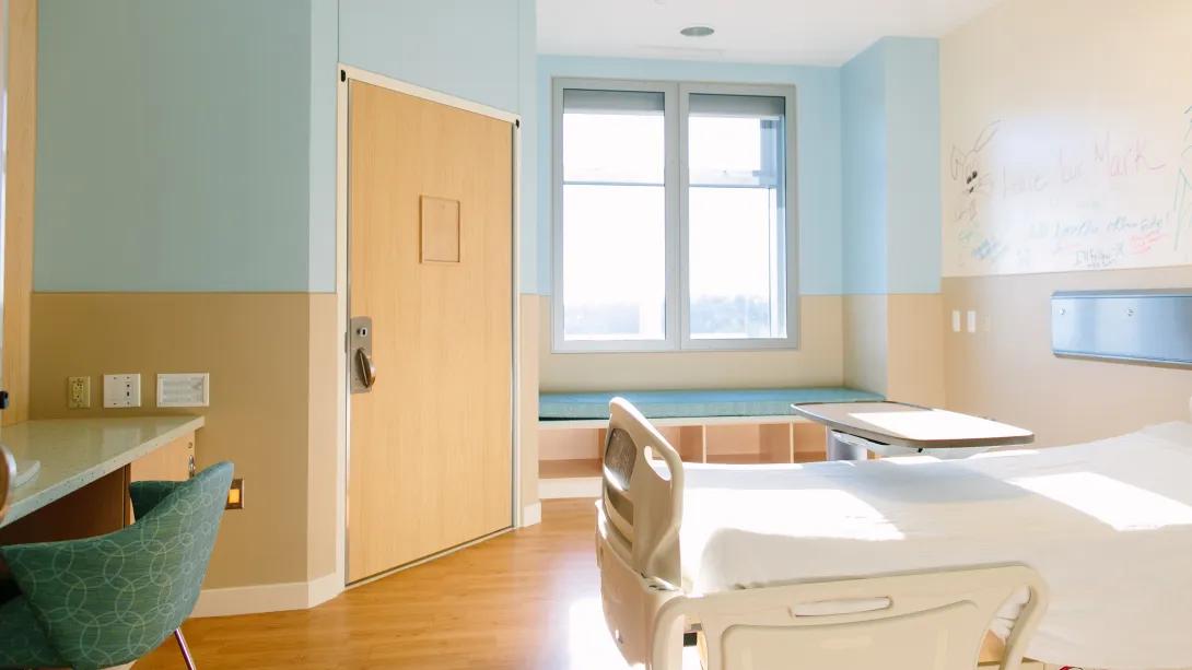 empty patient room with a bed, desk, window seat, and private bathroom