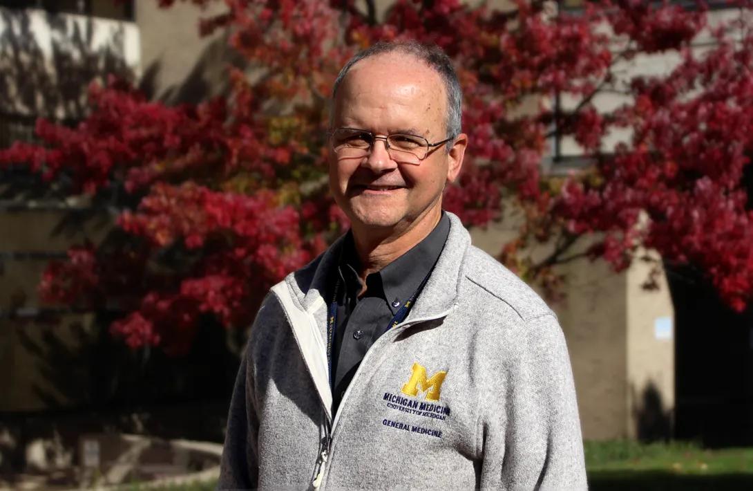 Man wearing glass and a grey Michigan zip-up sweater standing in front of red-leafed trees