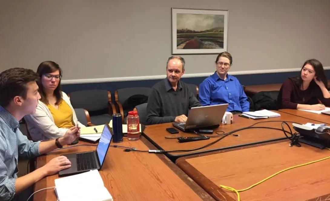 Five people sitting around a table, some with their laptops open