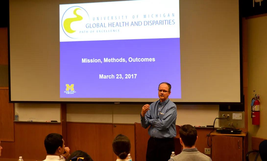 A man in front of a large screen giving a talk to people in a lecture hall