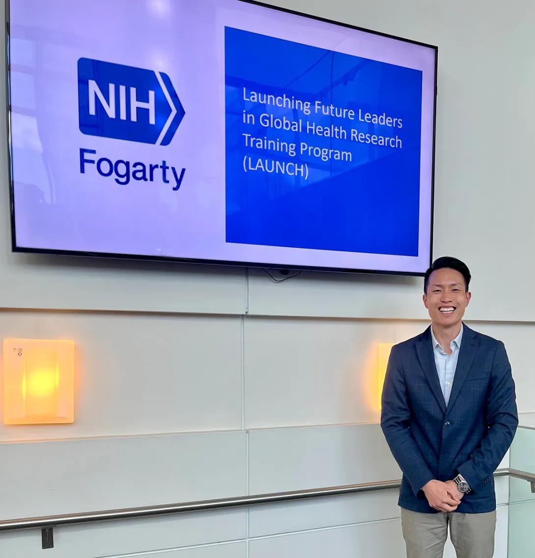 Man in a blazer standing next to a screen with a slide that reads 'NIH Fogarty'