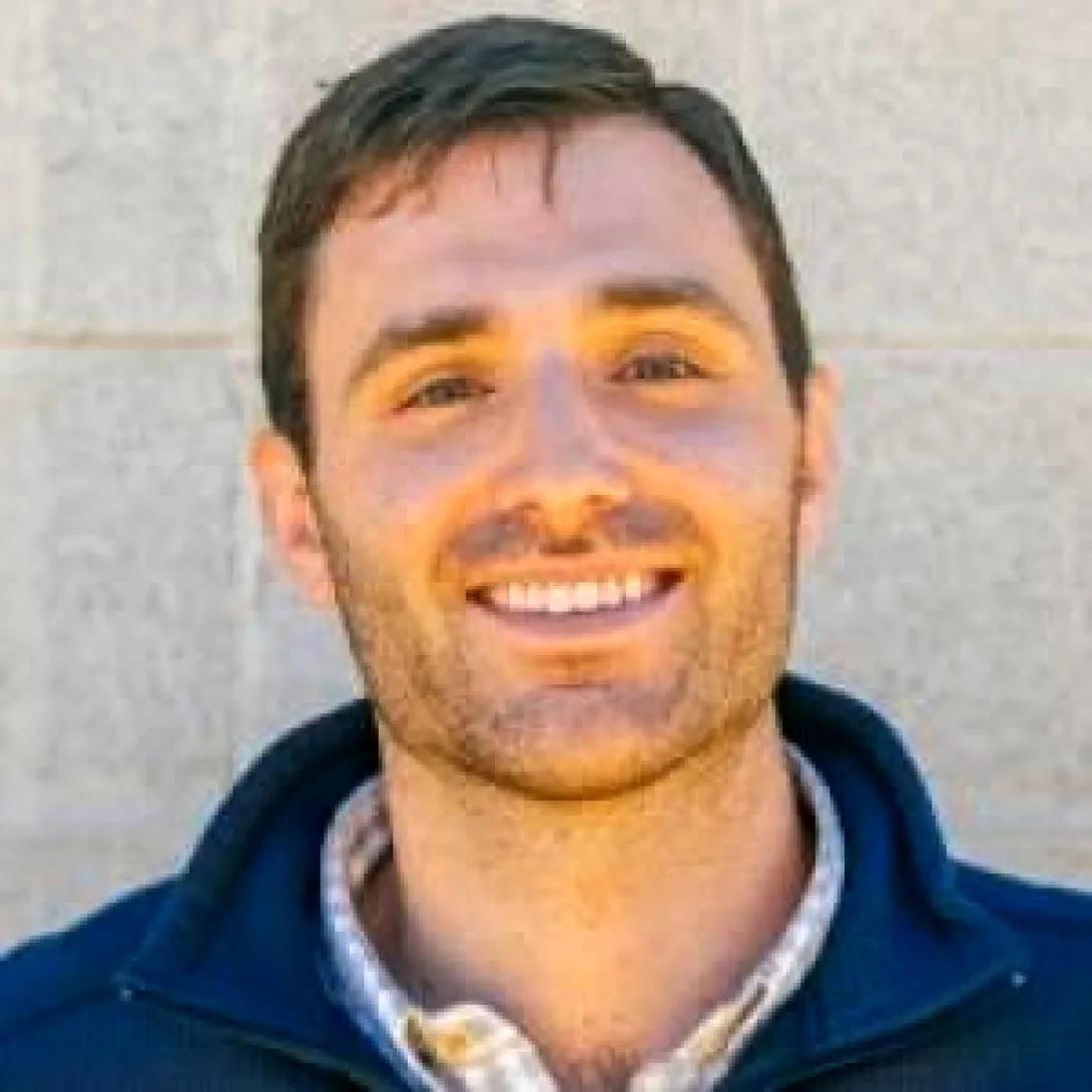 Man with short brown hair wearing a blue sweater over a white checked shirt