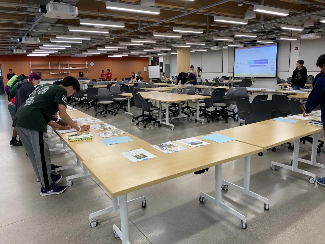a group of students stand at forestry stations at tables in a large room