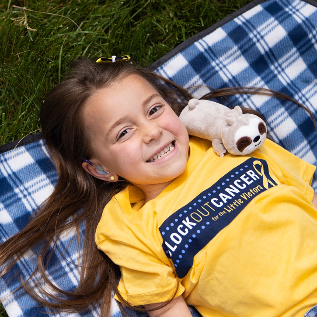 Smiling portrait of a girl in a Block Out Cancer t-shirt lying on a plaid blanket on a lawn. A plush sloth is leaned against her shoulder.
