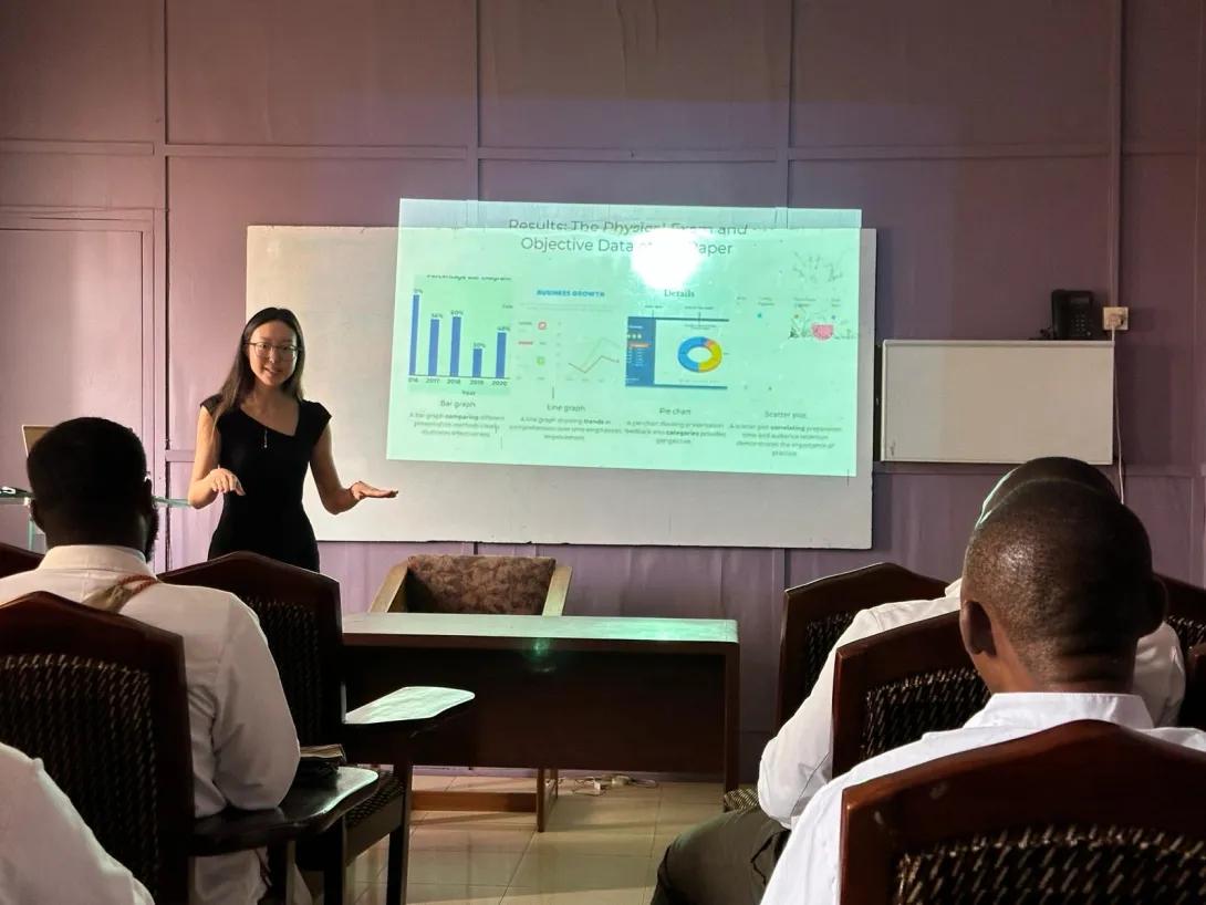 A woman speaks and shares a presentation on a screen to an audience in a classroom sized room