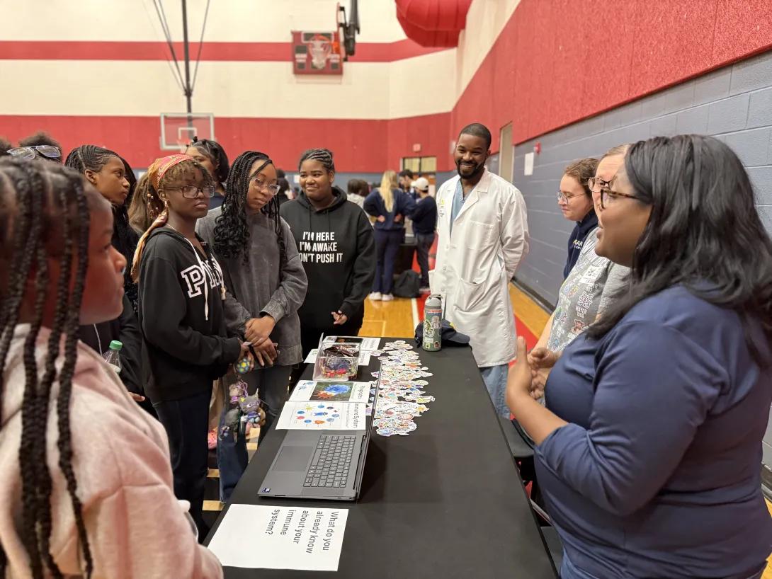 Group of students in a gym gather around a table where several doctors and medical professionals are talking