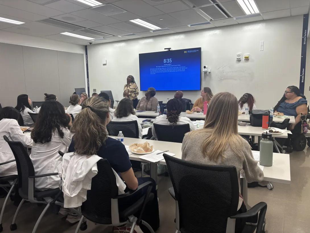 A panel of standardized patients sit at the front of a classroom while a group of medical students listen to their presentation
