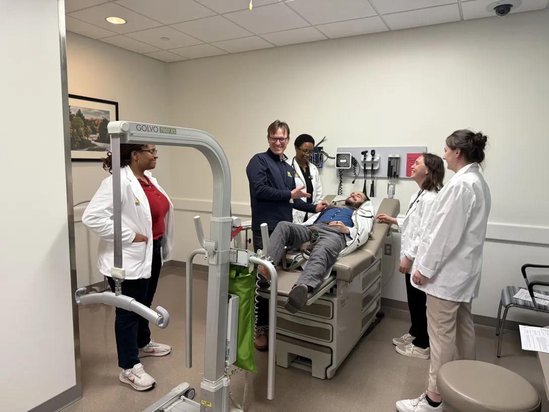 A group of four medical students in white coats with a faculty member learning to lift a patient on an exam table