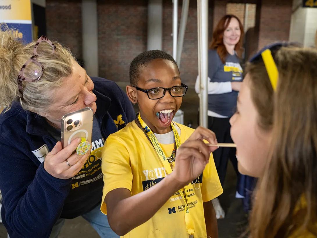 Middle school student smiles while administering a tongue depressor to another students tongue while an adult with a phone looks on.