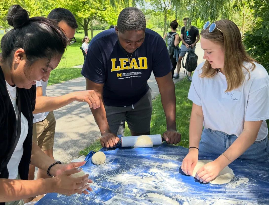 A groups of four medical students roll dough on a floured surface outside