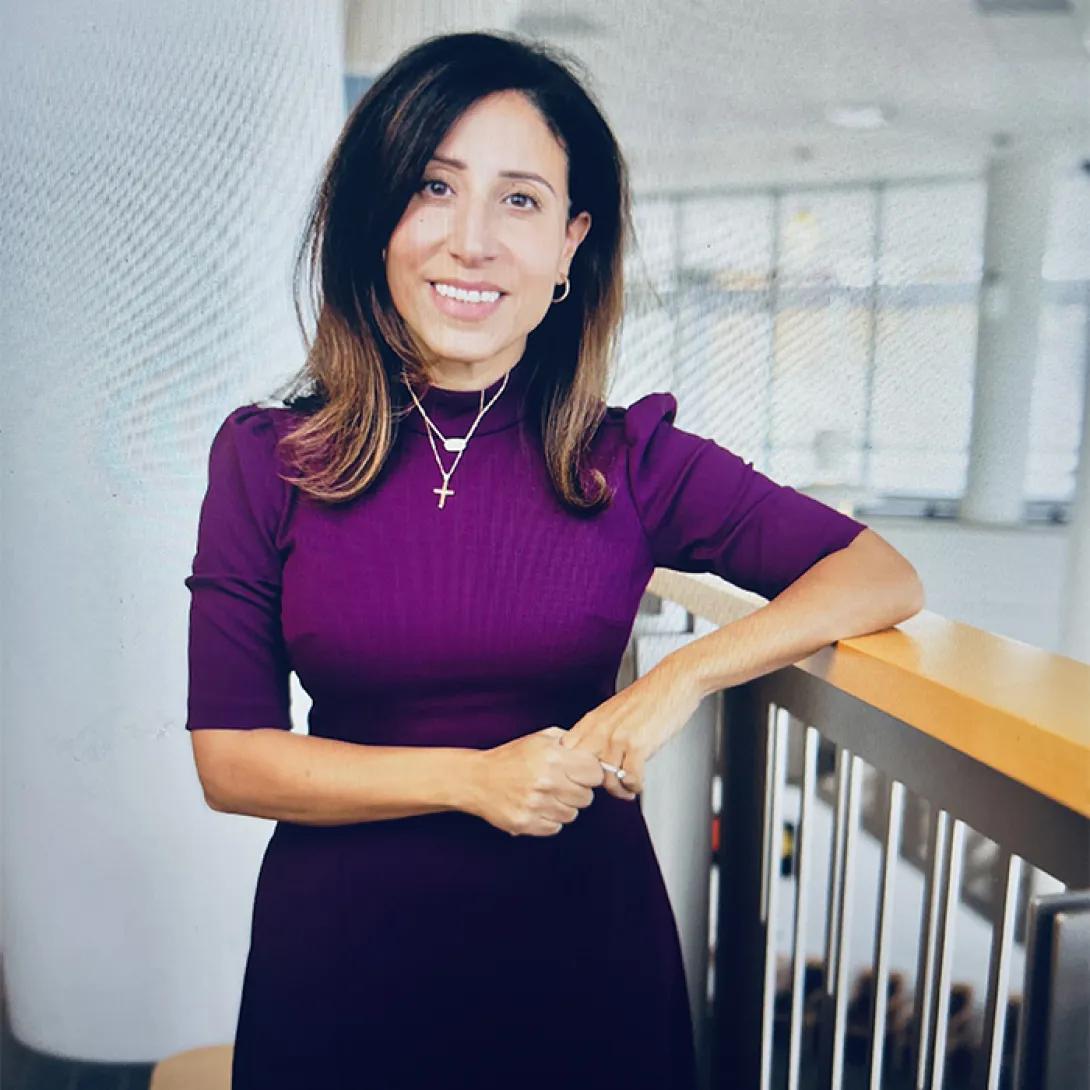 Julie Ishak, head and torso shot of white woman with shoulder-length brown hair wearing purple short-sleeved sweater and a cross necklace, hands folded at waist
