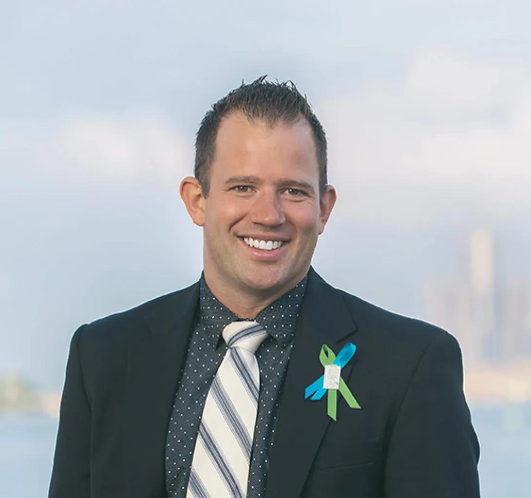 Smiling headshot of Luke Sparkman. He has blue and green awareness ribbons pinned to his lapel and is wearing a dark suit jacket.