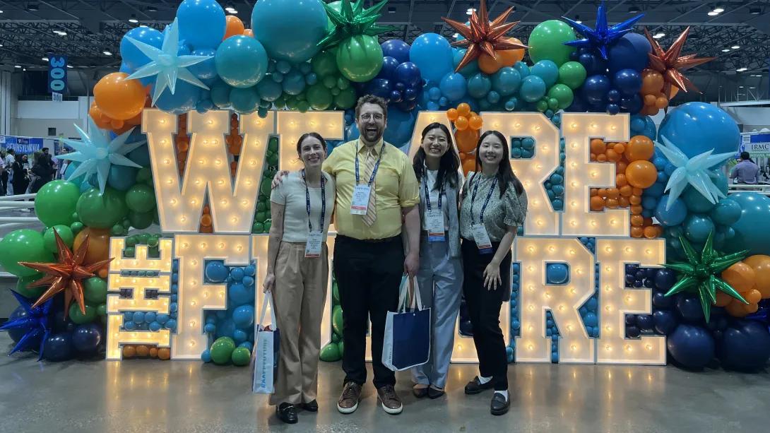 Four people stand together in front of a large illuminated "WE ARE THE FUTURE" sign, surrounded by colorful balloons and star decorations at a convention or event.
