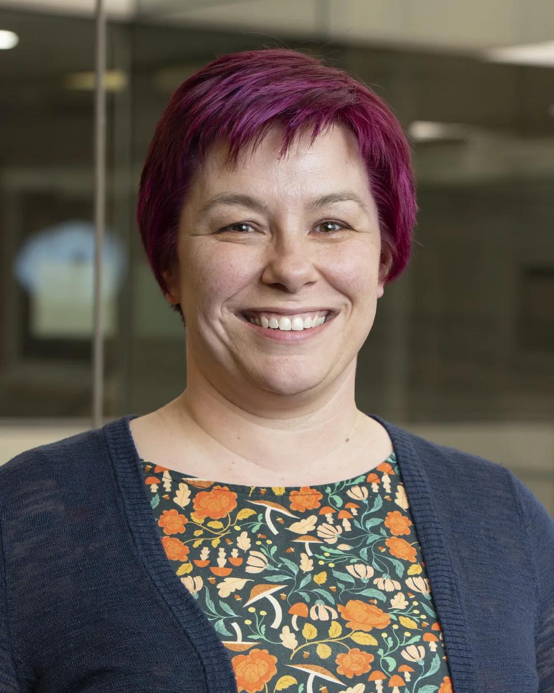 Head-and-shoulders portrait of a person with short magenta hair, wearing a navy cardigan over a dark floral top, standing indoors with a blurred background.