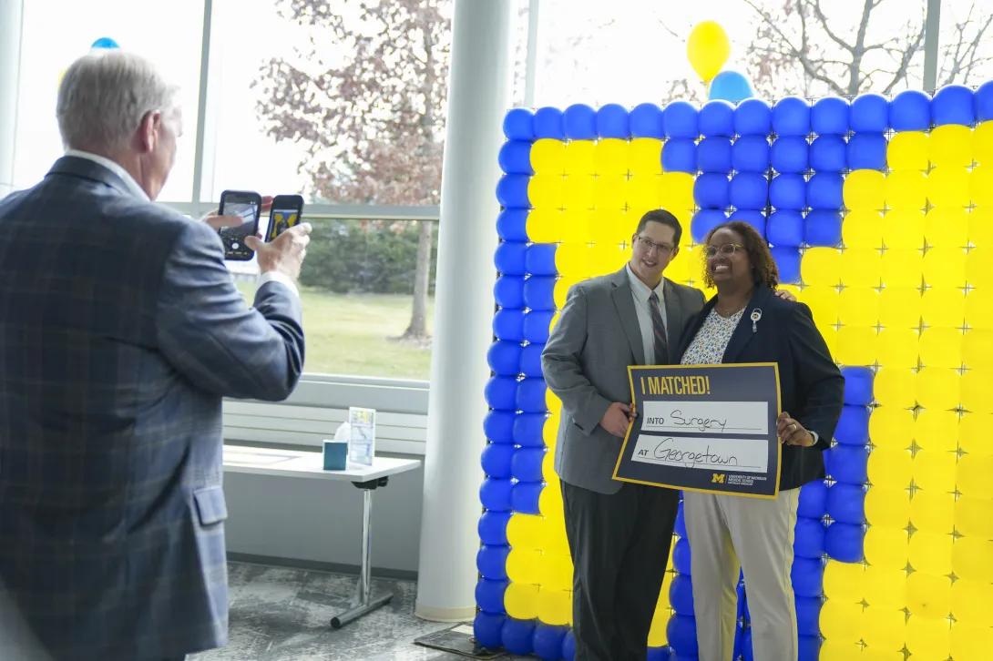 A person takes a photo of two people posing in front of a large maize-and-blue balloon backdrop while holding a University of Michigan Medical School “I MATCHED! Into Surgery at Georgetown” sign.